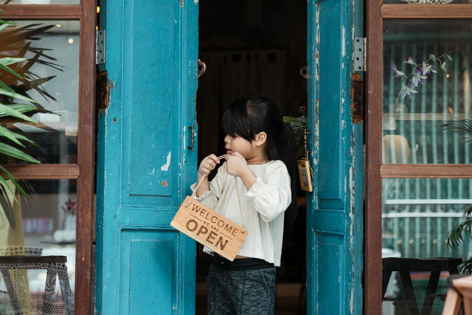 A young girl holding an open welcome sign while standing in a rustic doorway with blue doors.